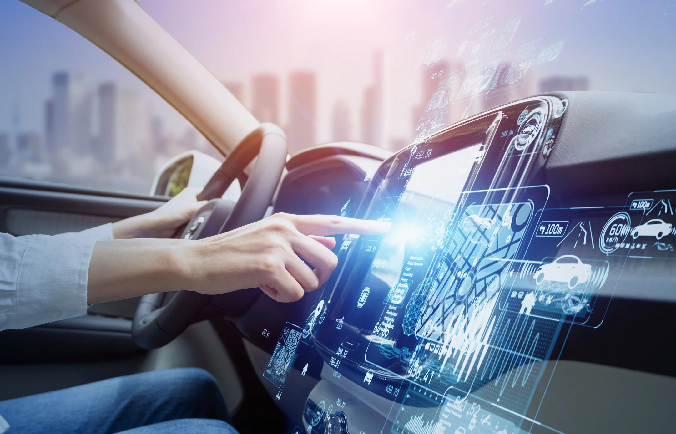 Person driving car with city in the background. Person is pointing to high-tech interface on the console.